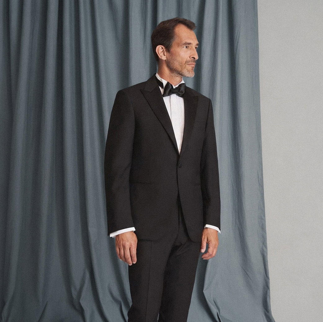 Mann im schwarzen Smoking mit weißem Hemd und schwarzer Fliege steht seitlich vor graublauem Vorhang. Mann im schwarzen Smoking mit weißem Hemd und schwarzer Fliege steht seitlich vor graublauem Vorhang.