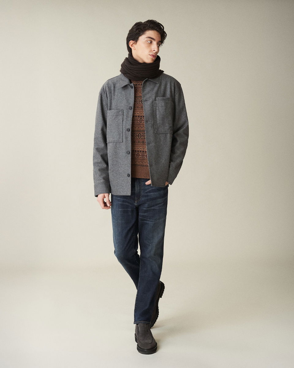 Mann trägt dunkle Jeans, braunen gemusterten Pullover, graues Overshirt und schwarzen Schal mit Doppelumwicklung, steht vor hellem Hintergrund.