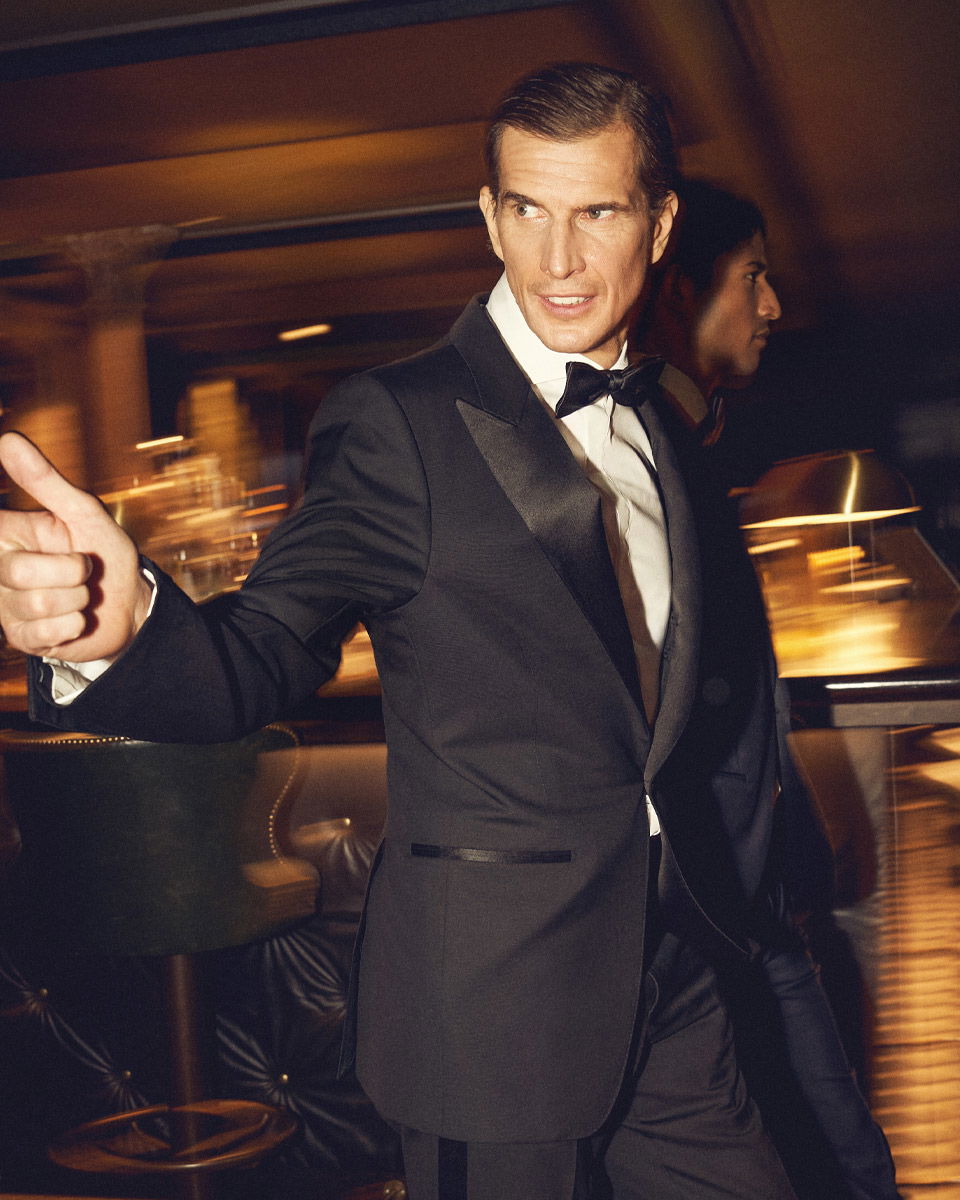 Mann im schwarzen Smoking mit weißem Hemd und schwarzer Fliege zeigt Daumen hoch in festlicher, eleganter Bar-Atmosphäre. Mann im schwarzen Smoking mit weißem Hemd und schwarzer Fliege zeigt Daumen hoch in festlicher, eleganter Bar-Atmosphäre.