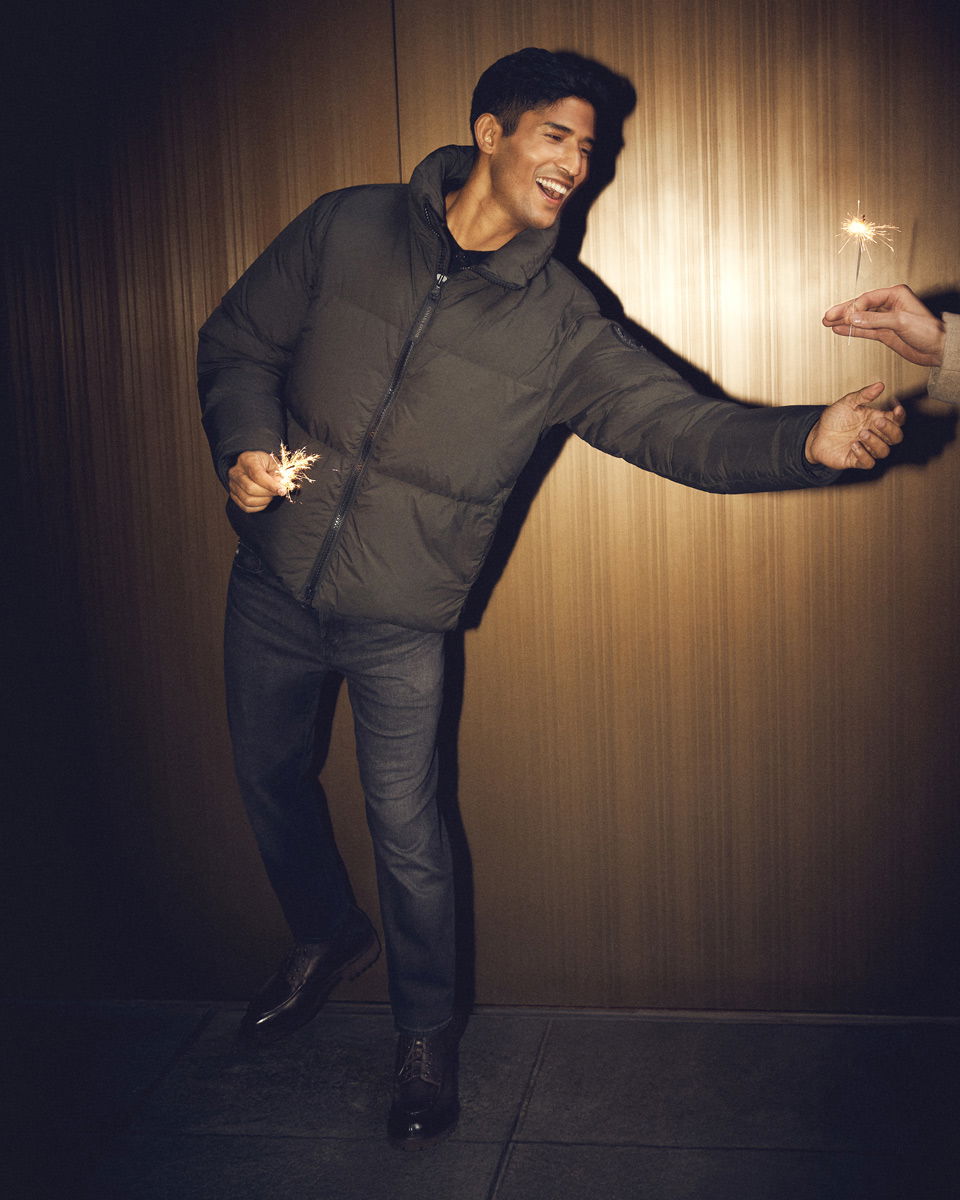 Junger Mann in dunkelgrauer Daunenjacke, Jeans und schwarzen Schuhen lacht und hält Wunderkerzen in der Hand. Junger Mann in dunkelgrauer Daunenjacke, Jeans und schwarzen Schuhen lacht und hält Wunderkerzen in der Hand.