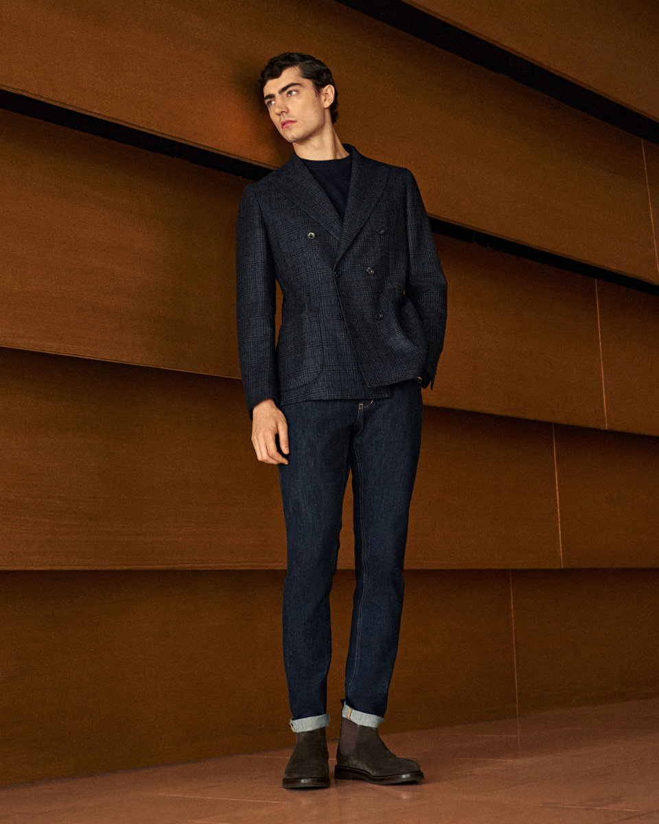 Mann trägt karierten dunkelblauen Zweireiher, navy Rundhalsshirt, dunkle Jeans mit Umschlag und graue Chelsea-Boots vor Holzwand.