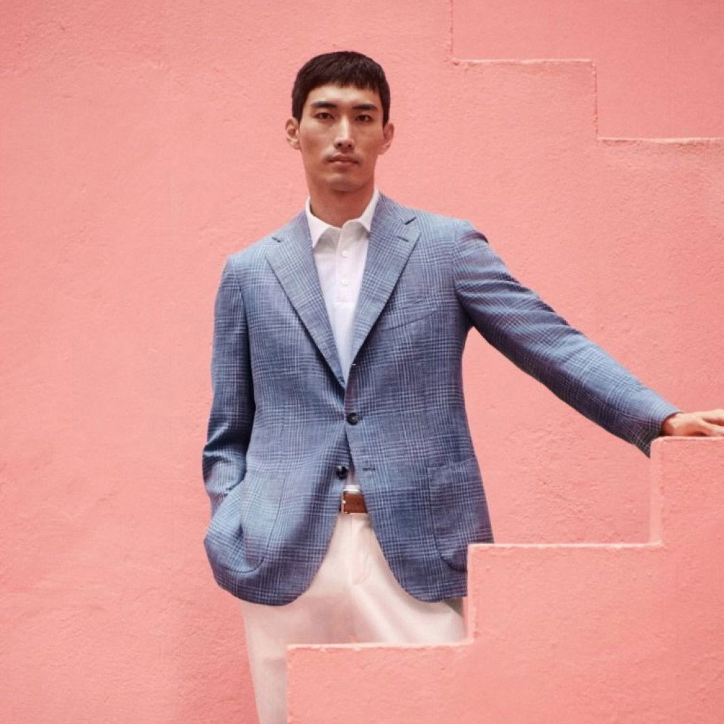 Mann mit kurzem schwarzen Haar trägt kariertes blau-weißes Sakko, weißes Hemd und cremefarbene Hose vor rosa Treppe.