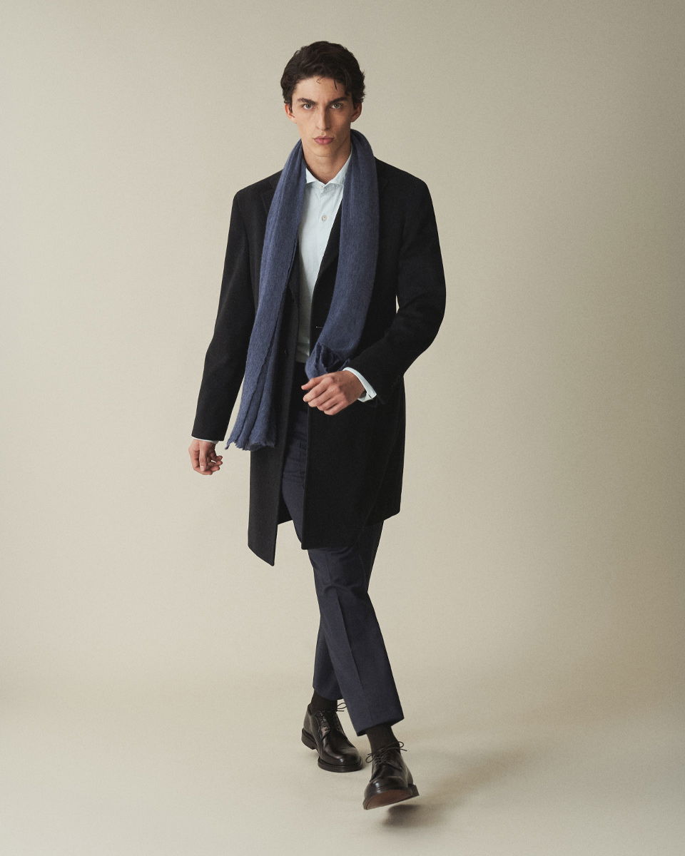 Mann in Bewegung trägt schwarzen Mantel, dunkelblauen Anzug, hellblaues Hemd, blauen Schal und braune Schnürschuhe.