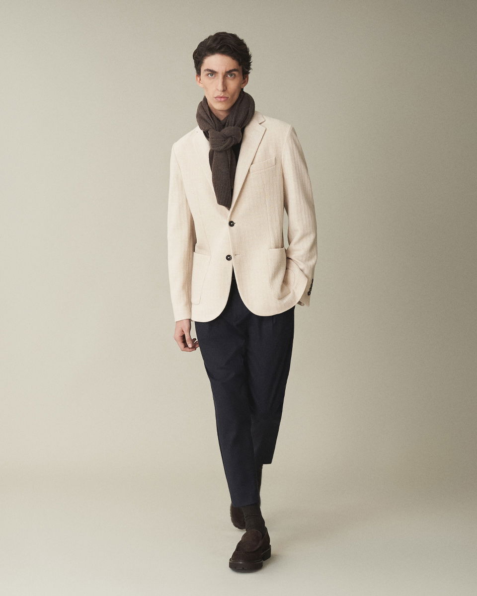 Mann mit beigem Sakko, schwarzer Hose, braunem Schal und braunen Loafers geht vor neutralem Hintergrund.