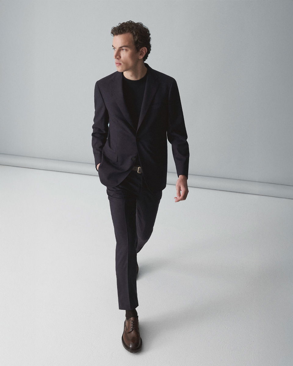 Mann in dunkelblauem Anzug, schwarzem Rundhalspullover und braunen Schnürschuhen geht selbstbewusst in hellem Raum. Mann in dunkelblauem Anzug, schwarzem Rundhalspullover und braunen Schnürschuhen geht selbstbewusst in hellem Raum.