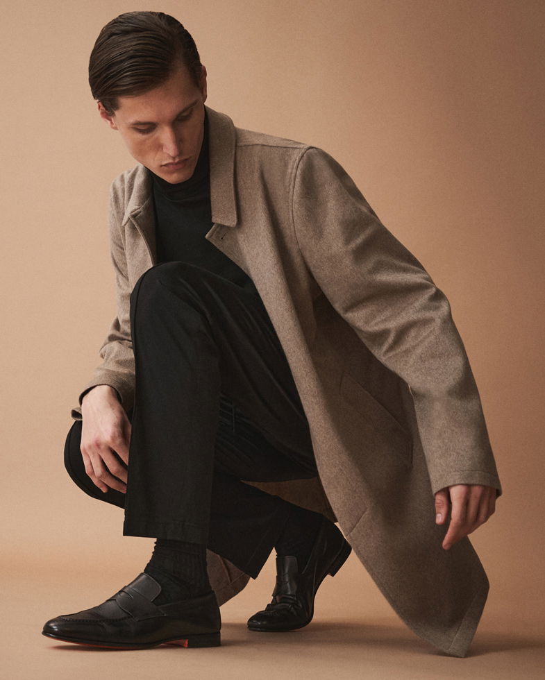 Mann kniet, trägt schwarzen Pullover, schwarze Hose, braunen Mantel und schwarze Loafer vor beige Hintergrund.