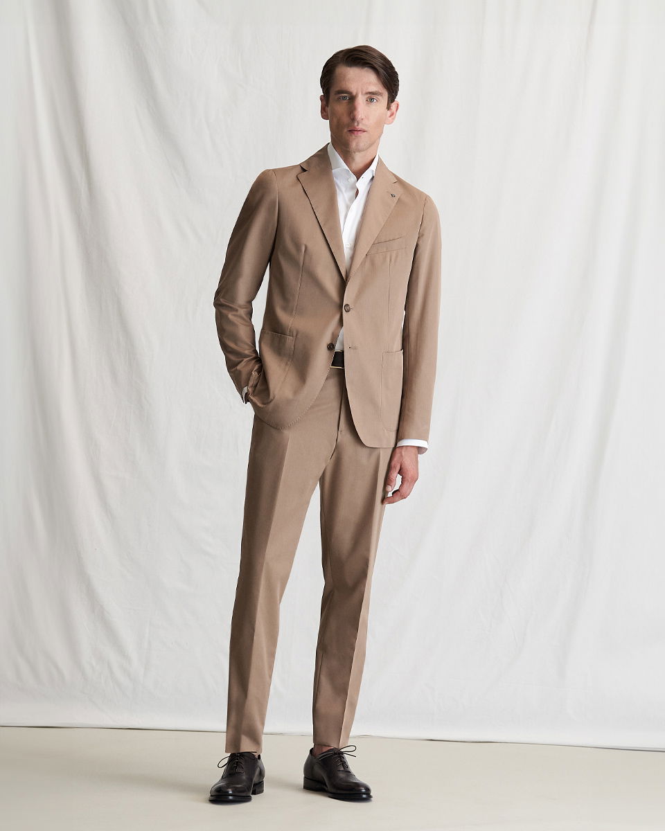 Mann steht vor hellem Hintergrund, trägt beige Anzug, weißes Hemd, schwarzen Gürtel und dunkelbraune Schnürschuhe. Mann steht vor hellem Hintergrund, trägt beige Anzug, weißes Hemd, schwarzen Gürtel und dunkelbraune Schnürschuhe.