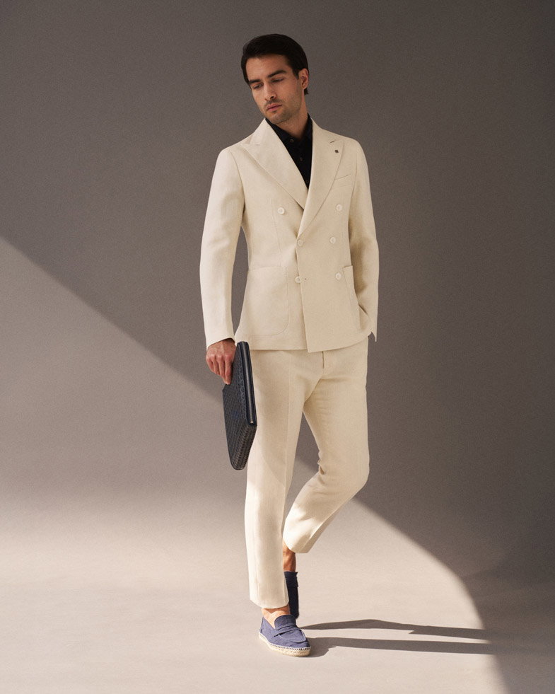 Eleganter Mann in cremefarbenem Anzug, schwarzem Hemd, dunkelblauen Espadrilles und schwarzer Aktentasche vor schlichtem Hintergrund. Eleganter Mann in cremefarbenem Anzug, schwarzem Hemd, dunkelblauen Espadrilles und schwarzer Aktentasche vor schlichtem Hintergrund.