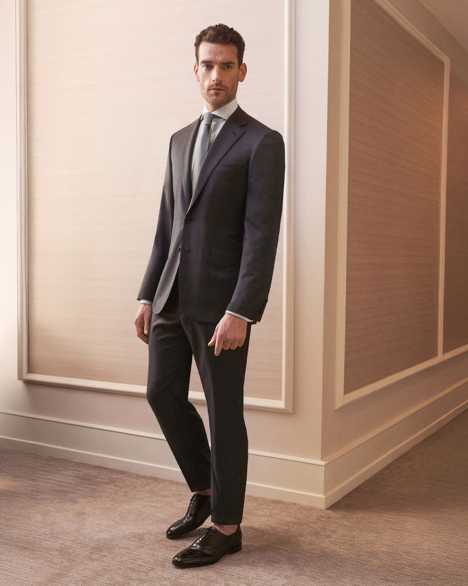 Mann in dunkelgrauem Anzug, hellgrauem Hemd, gemusterter Krawatte und schwarzen Schnürschuhen steht vor heller Wand.