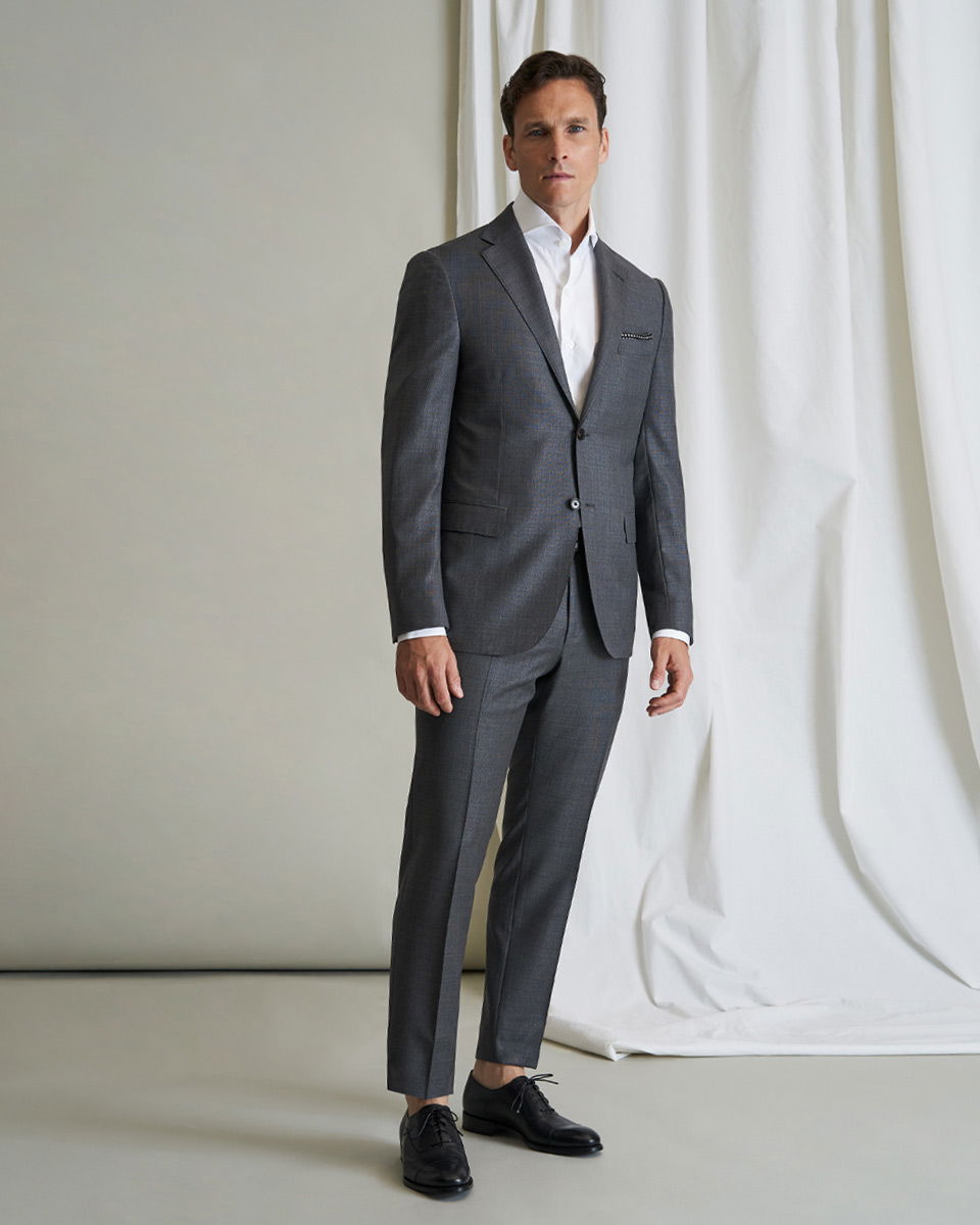 Mann in dunkelgrauem Anzug, weißem Hemd und schwarzen Schnürschuhen steht vor hellem Hintergrund mit weißem Vorhang.