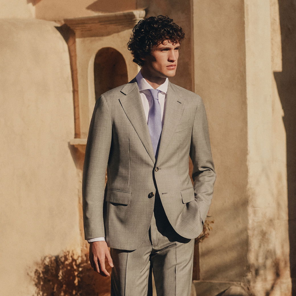 Junger Mann mit lockigem Haar trägt hellgrauen Anzug, weißes Hemd und fliederfarbene Krawatte, steht vor Sandsteinmauer. Junger Mann mit lockigem Haar trägt hellgrauen Anzug, weißes Hemd und fliederfarbene Krawatte, steht vor Sandsteinmauer.