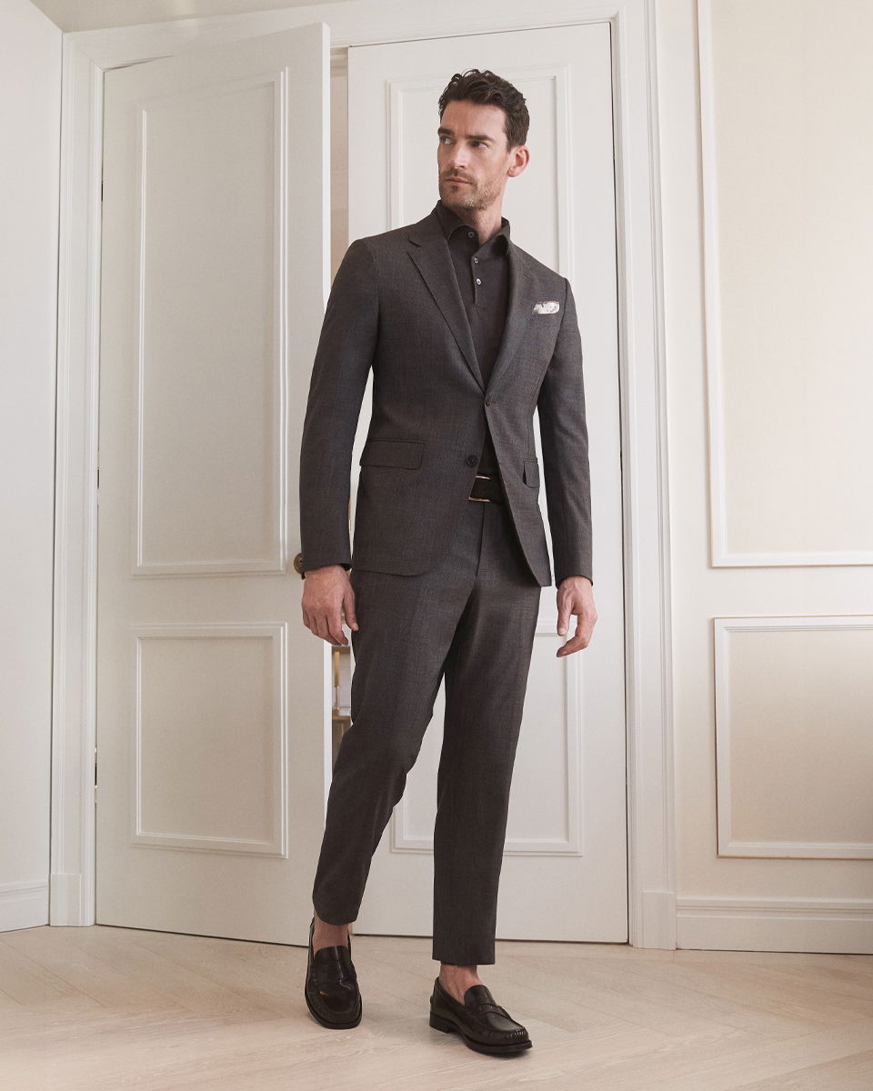 Mann trägt braunen Anzug mit dunklem Poloshirt, gemustertem Einstecktuch und schwarzen Loafern, steht vor weißer Tür.