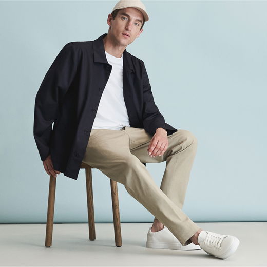 Mann mit beiger Baseballcap, weißem T-Shirt, blauem Overshirt, beiger Chinohose und weißen Sneakern sitzt auf Holzstuhl vor hellblauem Hintergrund.