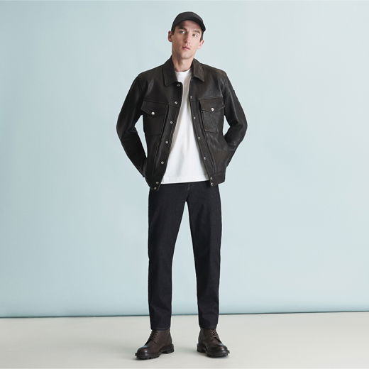 Mann mit schwarzer Baseball-Cap, schwarzer Lederjacke, weißem T-Shirt, schwarzer Hose und braunen Schnürboots vor hellblauem Hintergrund.