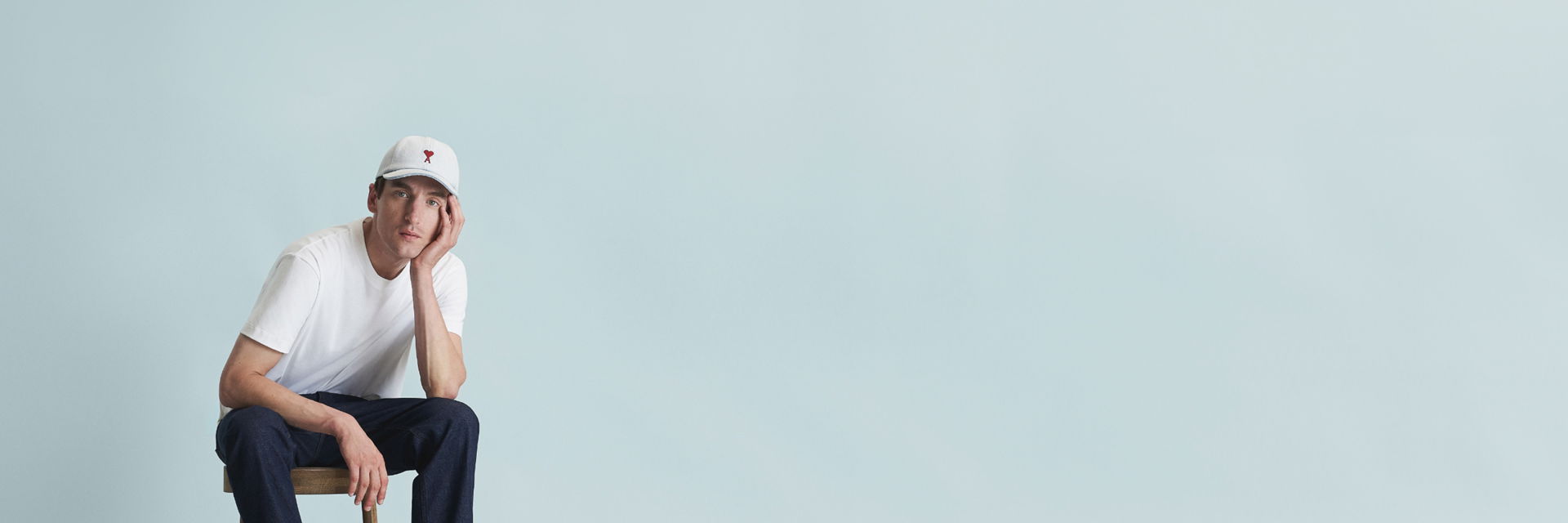 Mann sitzt auf Hocker, trägt weiße Baseball-Cap mit rotem Herzlogo, weißes T-Shirt und dunkelblaue Hose vor hellblauem Hintergrund. Mann sitzt auf Hocker, trägt weiße Baseball-Cap mit rotem Herzlogo, weißes T-Shirt und dunkelblaue Hose vor hellblauem Hintergrund.