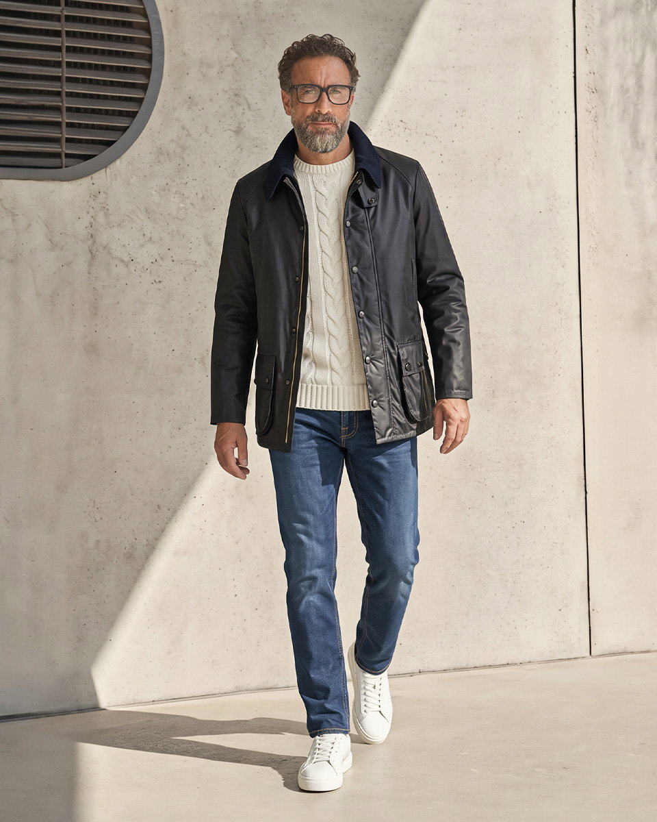 Mann mit Brille, weißem Strickpullover, schwarzer Wachsjacke, blauer Jeans und weißen Sneakern vor heller Wand.