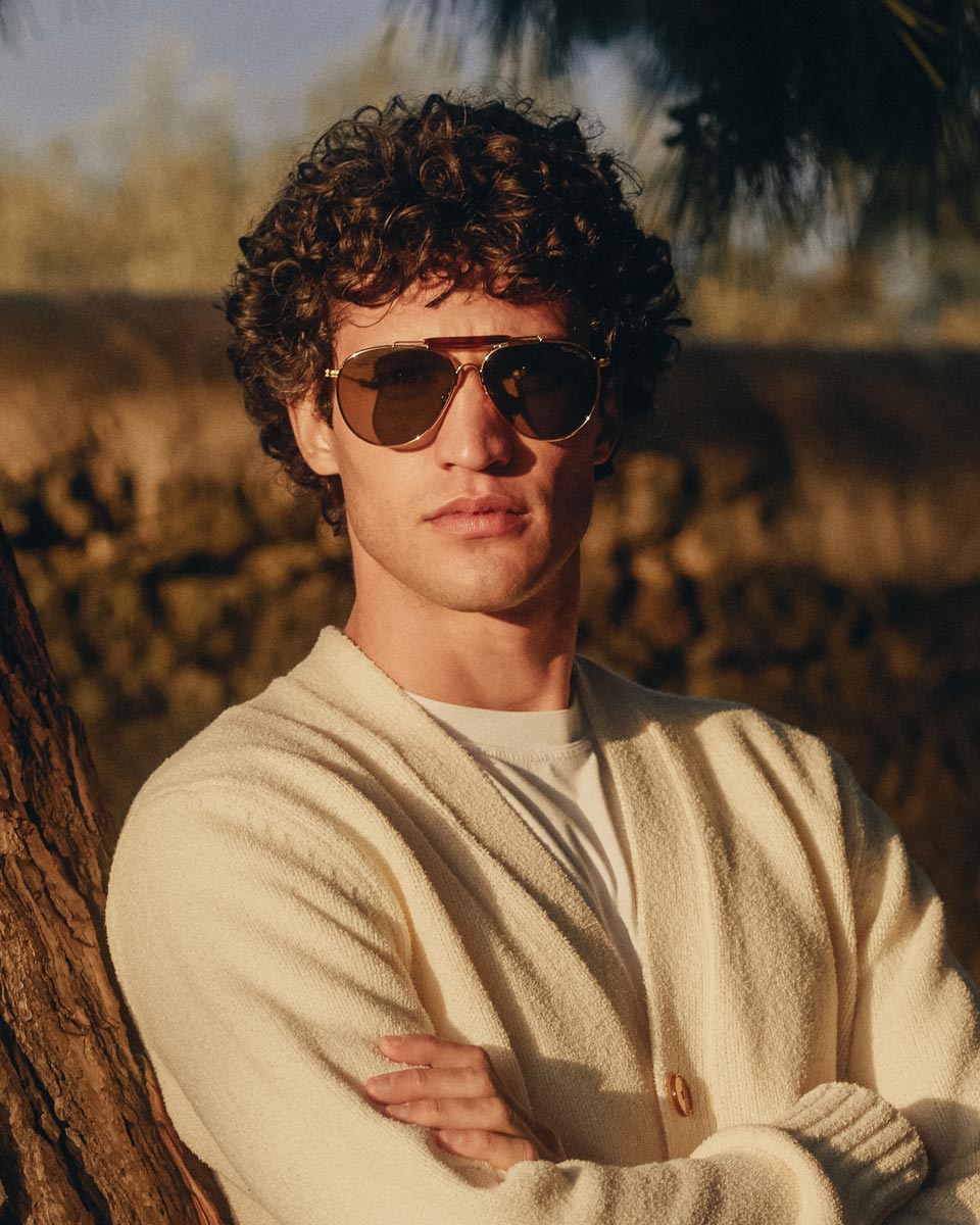 Junger Mann mit lockigem Haar, cremefarbenem Cardigan und brauner Pilotensonnenbrille mit goldenem Metallrahmen lehnt an Baum.