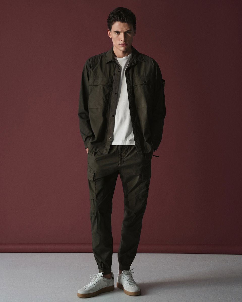 Mann steht vor rotem Hintergrund, trägt olivgrüne Cargohose, olivgrünes Overshirt, weißes T-Shirt und weiße Sneaker.