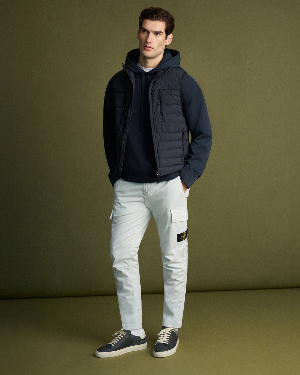 Mann trägt dunkelblauen Hoodie, schwarze gesteppte Weste, hellgraue Cargohose mit Logo-Patch und dunkelblaue Sneaker vor grünem Hintergrund.