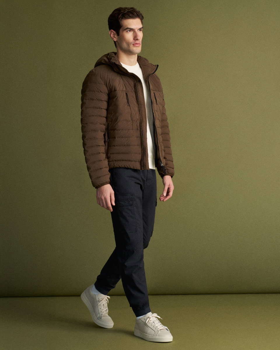 Mann trägt braune Steppjacke mit Kapuze, weißes Shirt, navyblaue Cargohose und weiße Sneaker vor grünem Hintergrund.