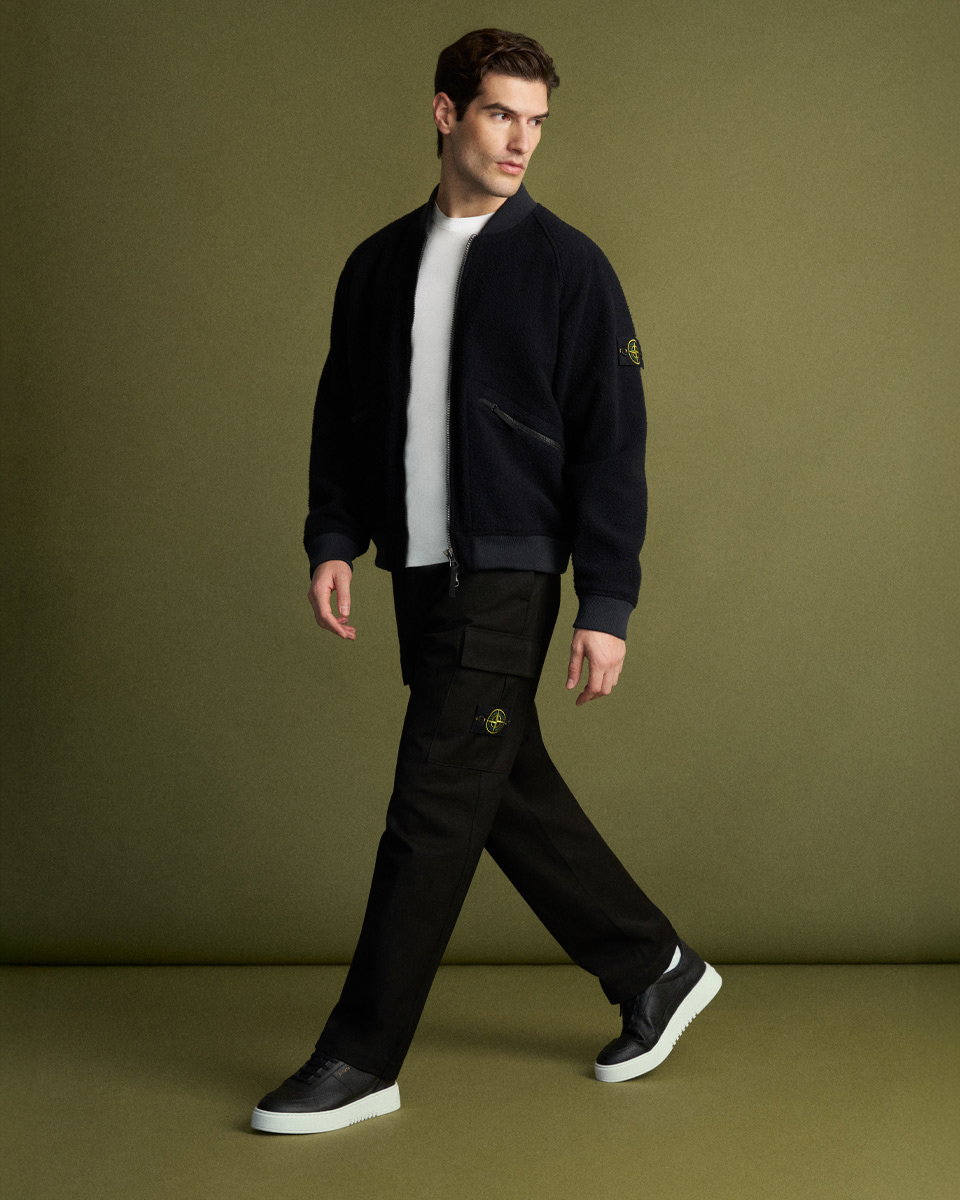 Mann trägt schwarze Cargohose, weißes T-Shirt, schwarzen Blouson mit Logo am Ärmel und schwarze Sneaker mit weißer Sohle.