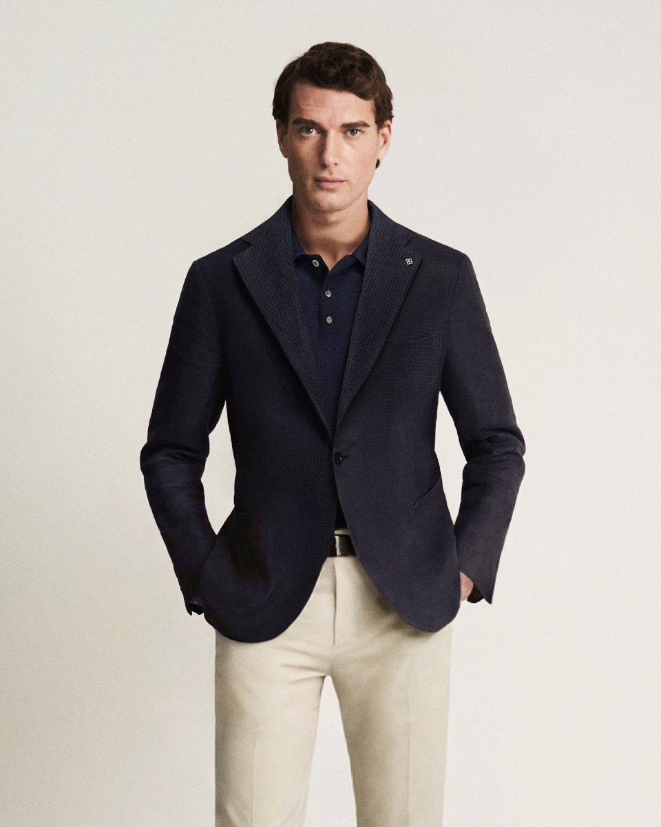 Mann trägt dunkelblaues Sakko, dunkelblaues Poloshirt und beige Chinohose, Hände in Taschen, neutraler Hintergrund.