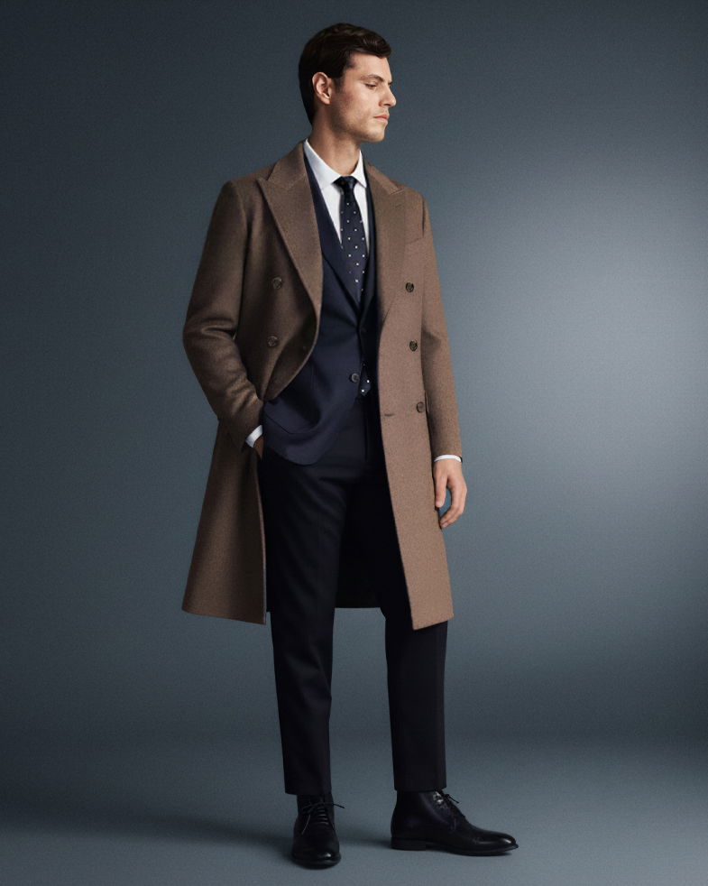 Mann trägt dunkelblauen Anzug, weißes Hemd, gepunktete Krawatte und braunen Mantel, steht seitlich vor grauem Hintergrund.