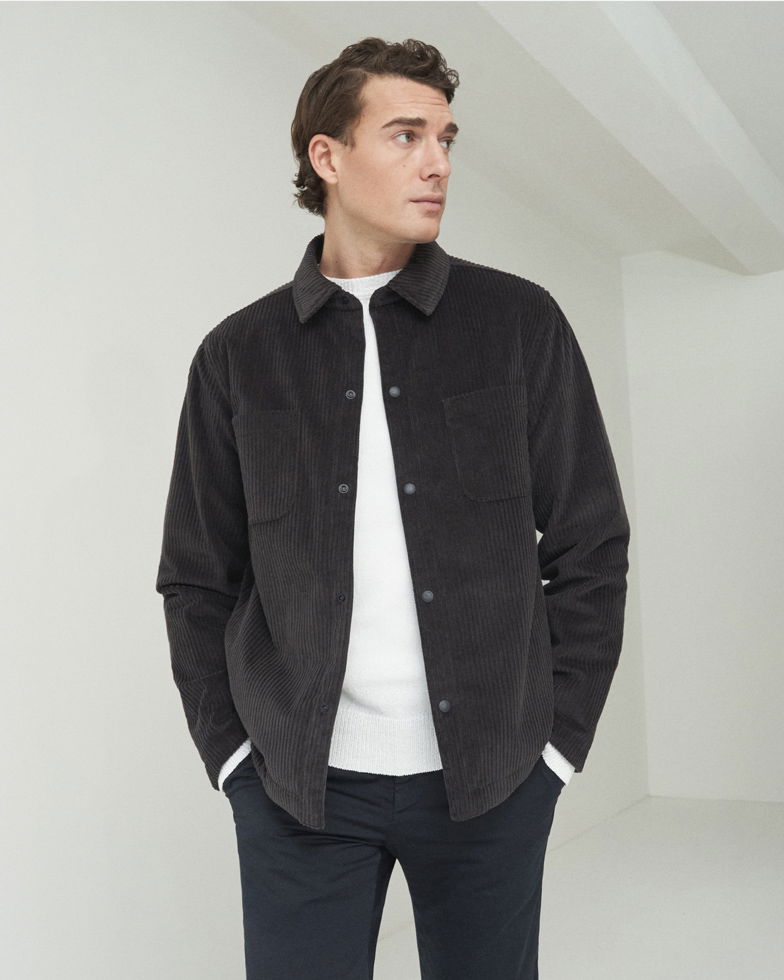 Mann trägt schwarzes Cord-Overshirt, weißes Strickshirt und dunkelblaue Hose, steht vor schlichtem, hellem Hintergrund.