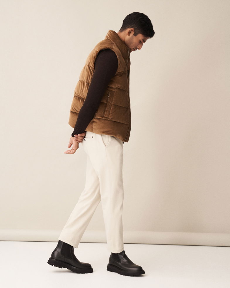 Mann in beige Cordhose, schwarzem Pullover, brauner Daunenweste und schwarzen Chelsea Boots vor hellem Hintergrund.
