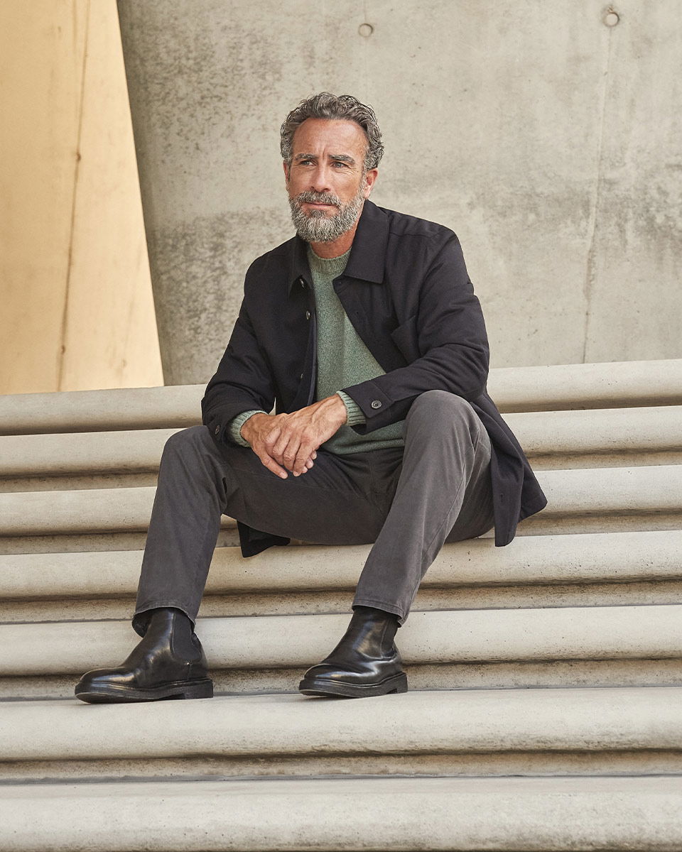 Mann mit grauem Pullover, dunkelgrauer Cordhose, schwarzem Mantel und schwarzen Chelsea Boots sitzt entspannt auf Treppen aus Beton. Mann mit grauem Pullover, dunkelgrauer Cordhose, schwarzem Mantel und schwarzen Chelsea Boots sitzt entspannt auf Treppen aus Beton.