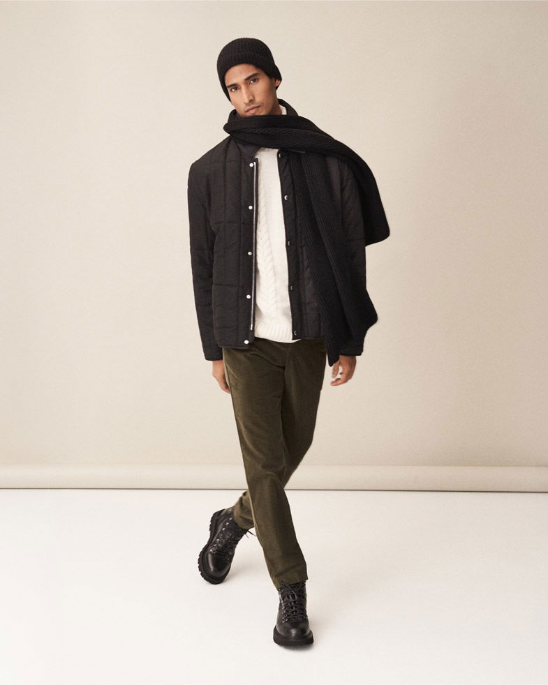 Mann trägt schwarze Daunenjacke, weißen Pullover, grünen Cordhose, schwarzen Schal, Mütze und braune Schnürboots, läuft.