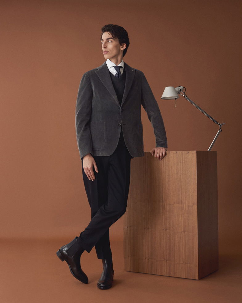 Mann steht vor braunem Hintergrund, trägt graues Cordsakko, weißes Hemd, schwarzen Pullover, schwarze Hose, Boots und blaue Krawatte.
