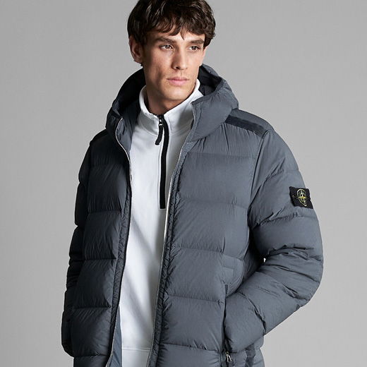 Mann mit braunen Haaren trägt graue Daunenjacke mit Kapuze und weißen Troyer mit Reißverschluss vor grauem Hintergrund.