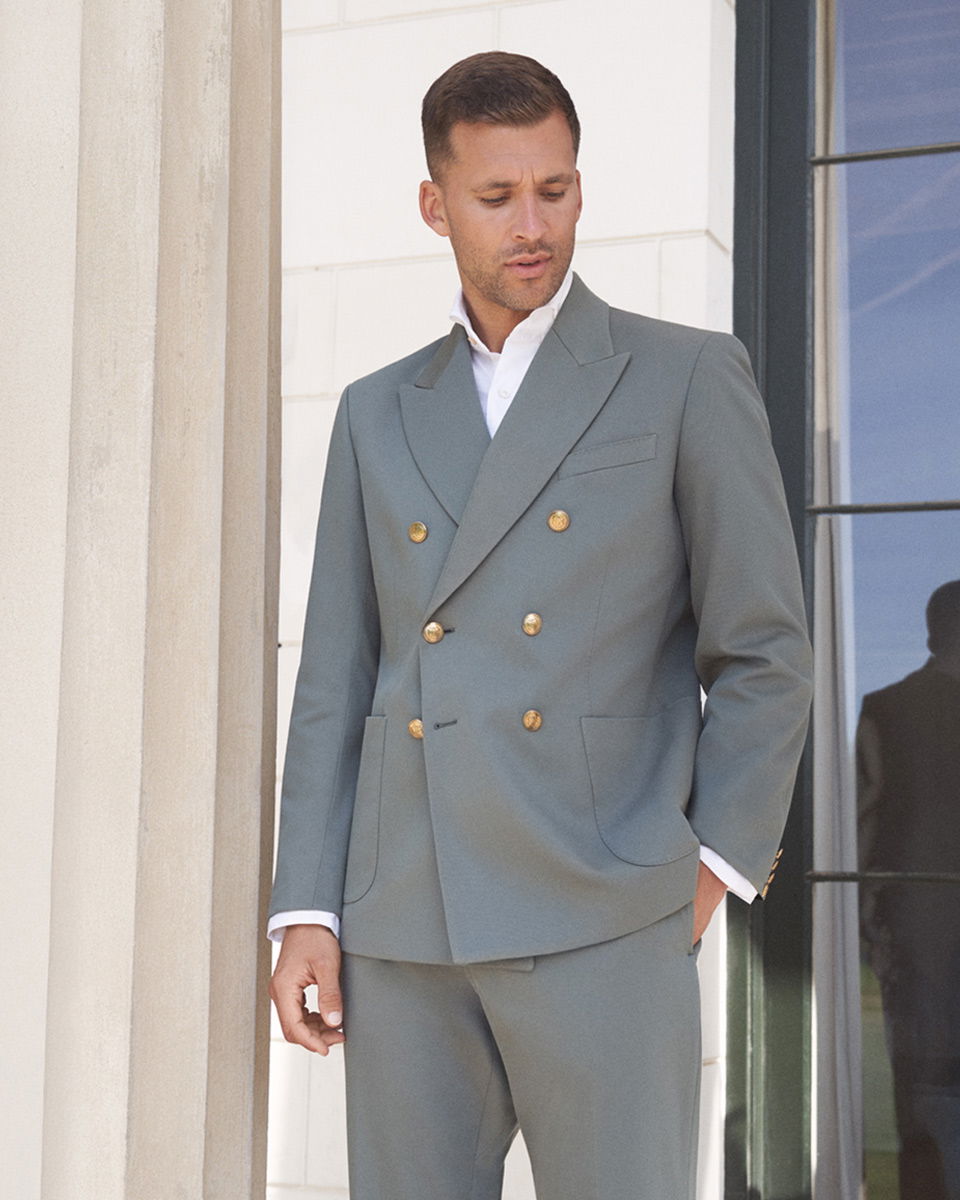 Mann trägt graugrünes Doppelreiher-Sakko mit goldenen Knöpfen und weiße Hemd, steht vor weißer Wand und Fenster.