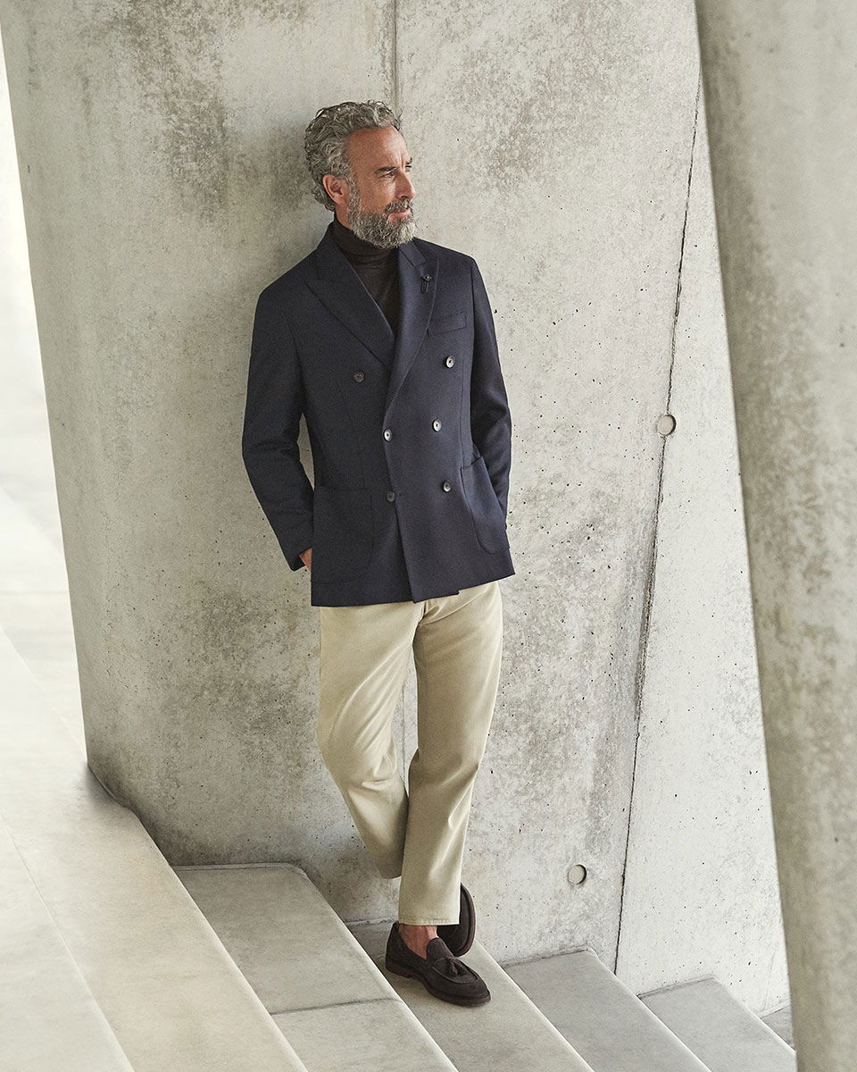 Herr mit grauem Haar und Bart trägt dunkelblauen Doppelreiher, schwarzen Rollkragenpullover, beige Hose und braune Loafer, lehnt an Betonwand. Herr mit grauem Haar und Bart trägt dunkelblauen Doppelreiher, schwarzen Rollkragenpullover, beige Hose und braune Loafer, lehnt an Betonwand.