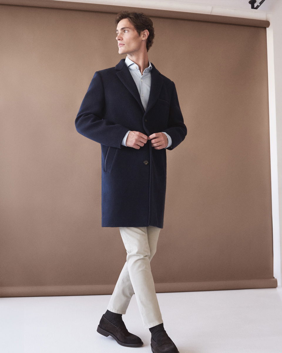 Mann trägt dunkelblauen Mantel, hellblaues Hemd, beige Hose und dunkelbraune Veloursleder-Loafer, steht vor braunem Hintergrund.
