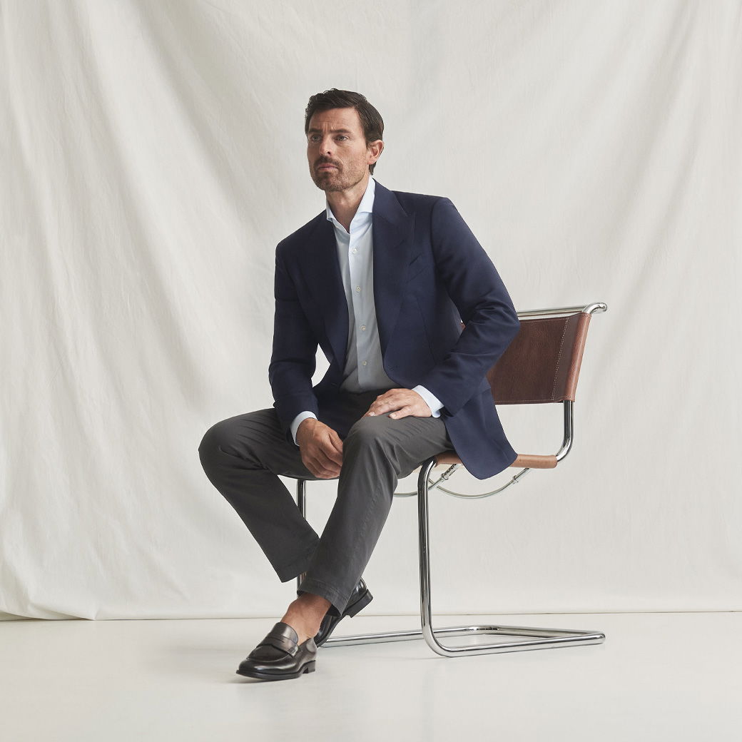 Mann sitzt auf Stuhl, trägt dunkelblaues Sakko, hellblaues Hemd, dunkelgraue Hose und schwarze Loafer, vor weißem Hintergrund.