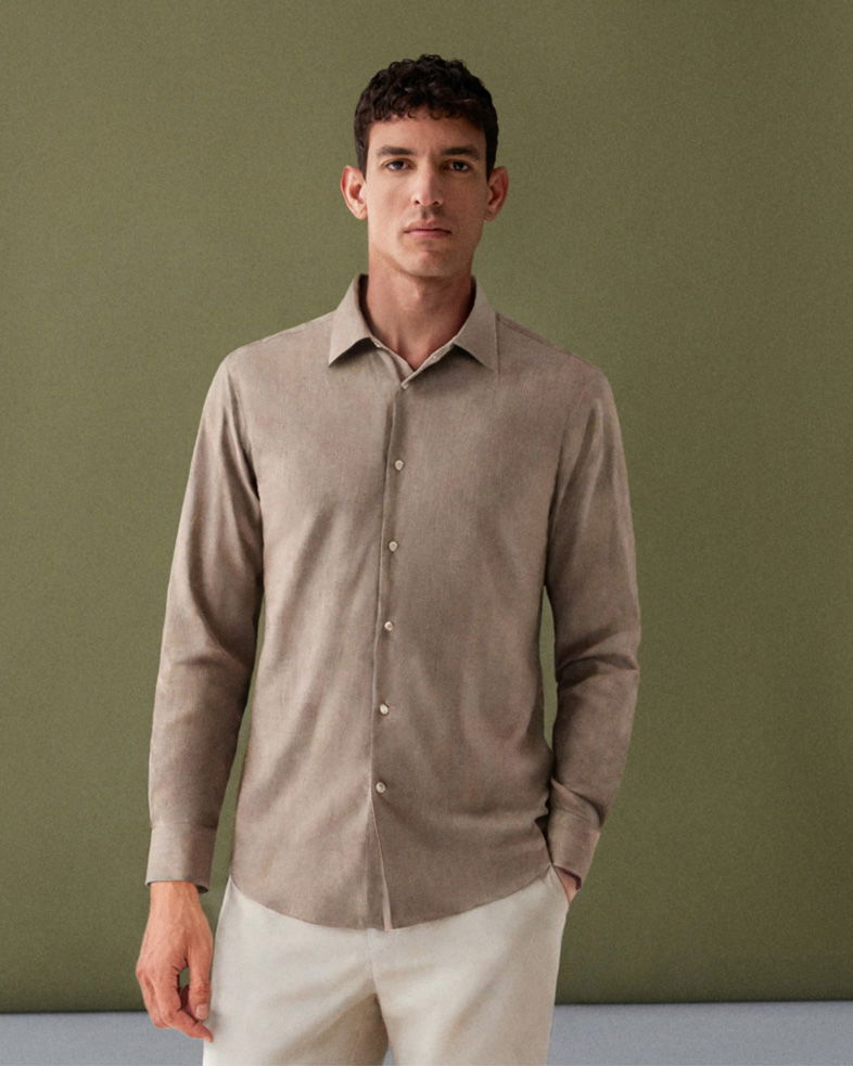 Mann mit kurzem dunklem Haar, trägt hellbraunes Hemd und cremefarbene Hose, steht vor grünem Hintergrund.