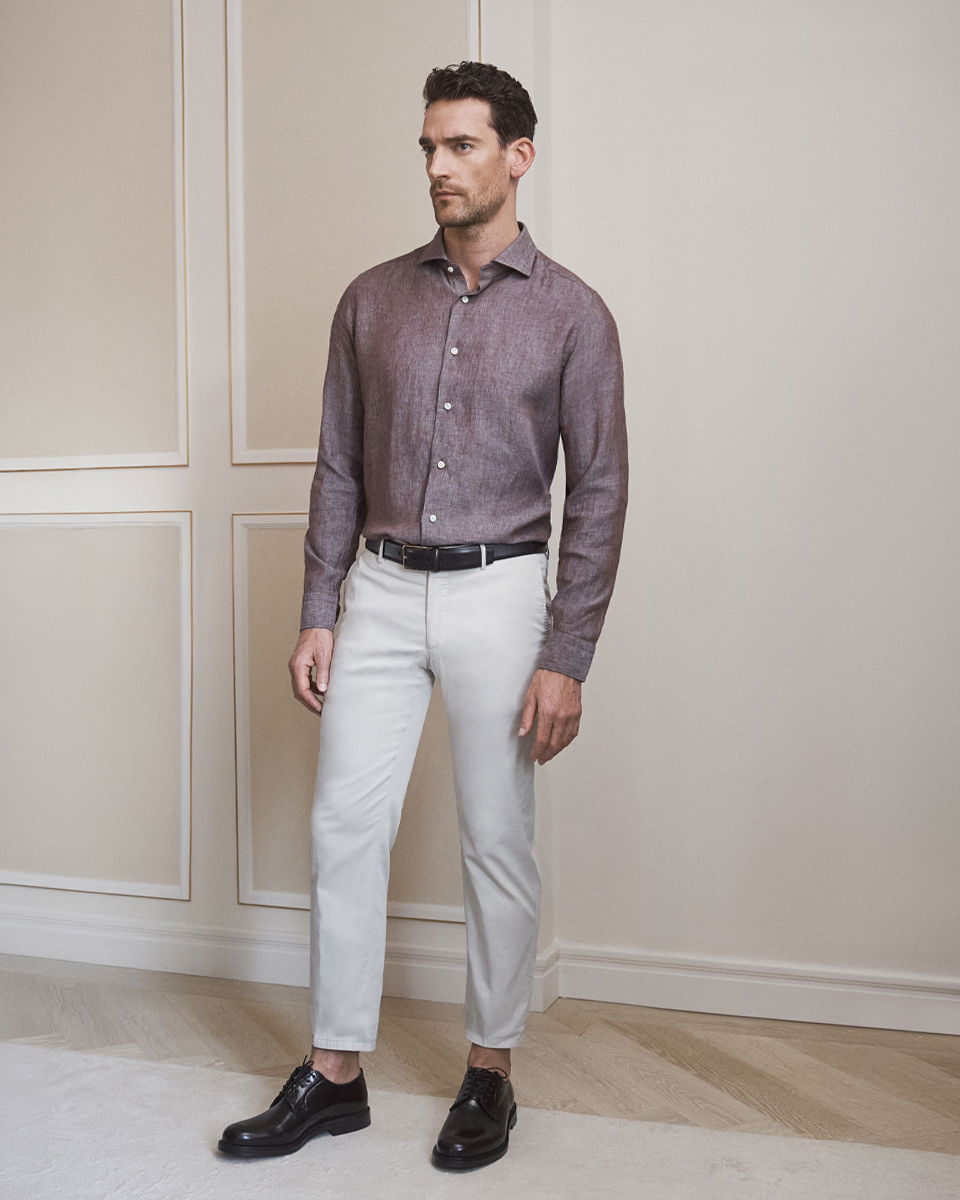 Mann trägt braun gemustertes Hemd, cremefarbene Chino, schwarzen Gürtel und schwarze Schnürschuhe, steht vor Wand mit Zierleisten.