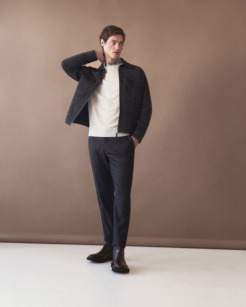 Mann trägt kariertes Hemd, weißen Pullover, dunkelblaue Hose, schwarze Velourslederjacke und schwarze Boots vor braunem Hintergrund.