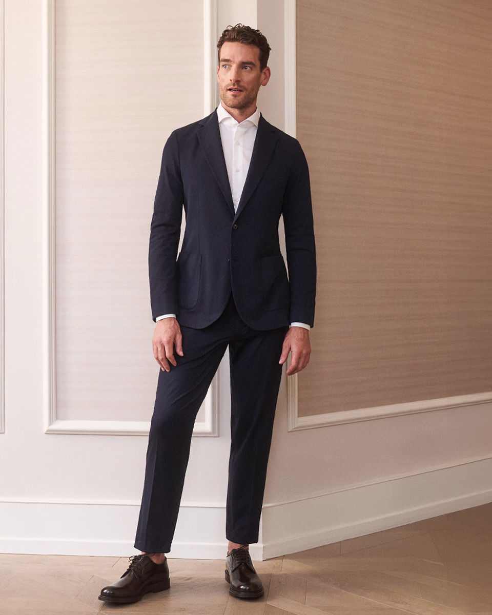 Mann in navy Anzug, weißem Hemd und braunen Lederschnürschuhen steht vor beige-weißem Wandhintergrund in elegantem Raum.