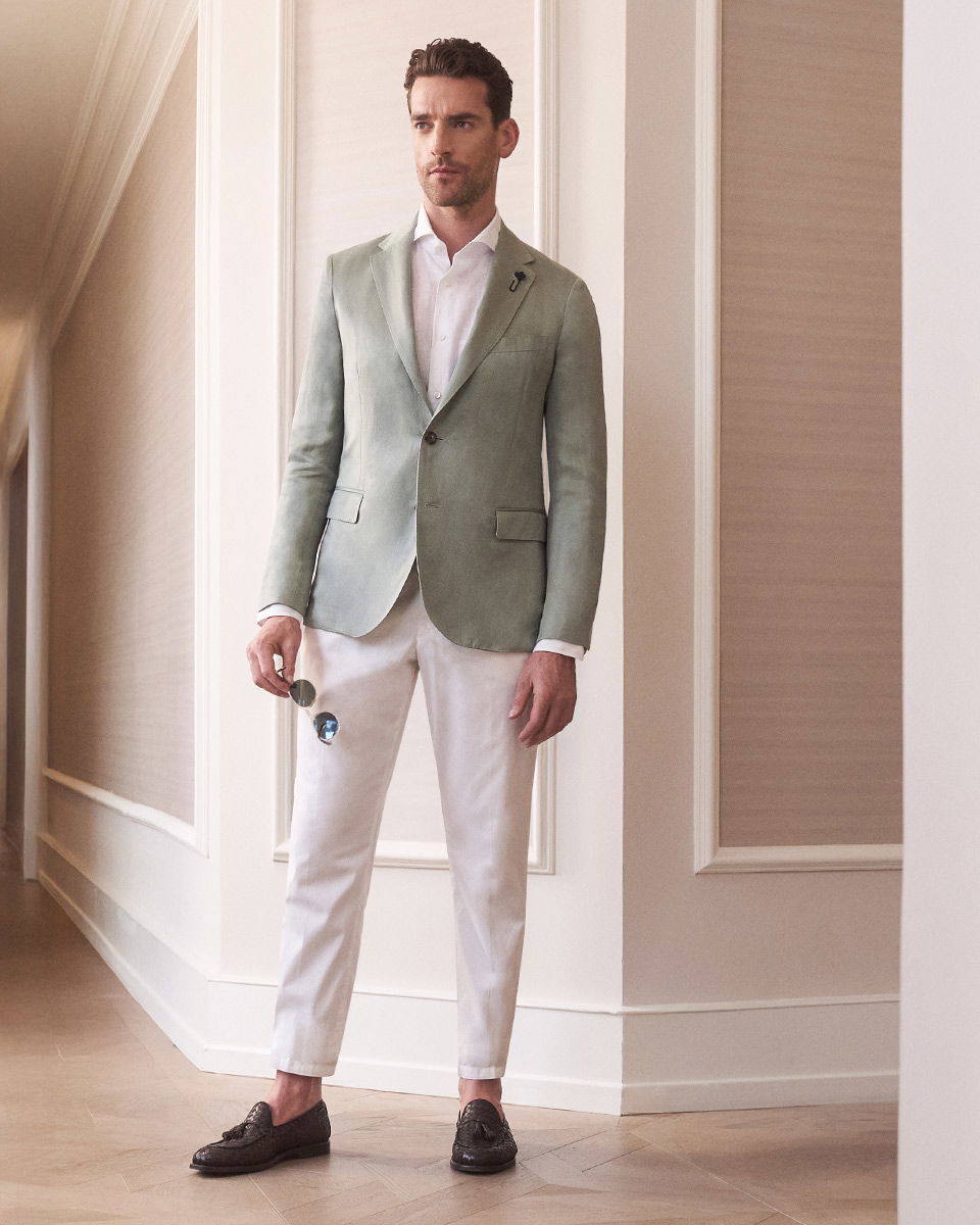 Mann trägt mintgrünes Sakko, weißes Hemd, weiße Hose, hält Sonnenbrille, dunkle Loafer, steht in elegantem Raum.