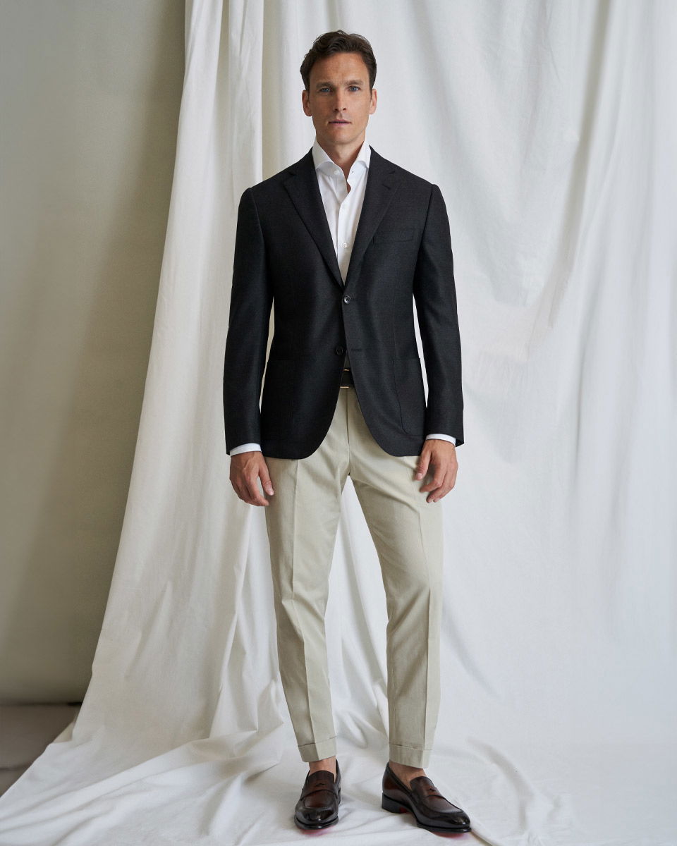 Mann trägt schwarzes Sakko, weißes Hemd, beige Chinohose und dunkelbraune Loafer, steht vor weißem Stoffhintergrund.