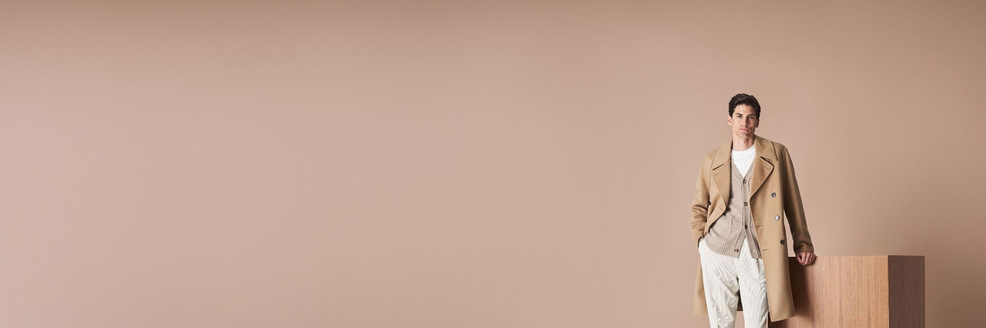 Mann lehnt an Holzblock, trägt beige Mantel, beigen Cardigan, weiße Hose, weißes Shirt und braune Boots, beiger Hintergrund. Mann lehnt an Holzblock, trägt beige Mantel, beigen Cardigan, weiße Hose, weißes Shirt und braune Boots, beiger Hintergrund.