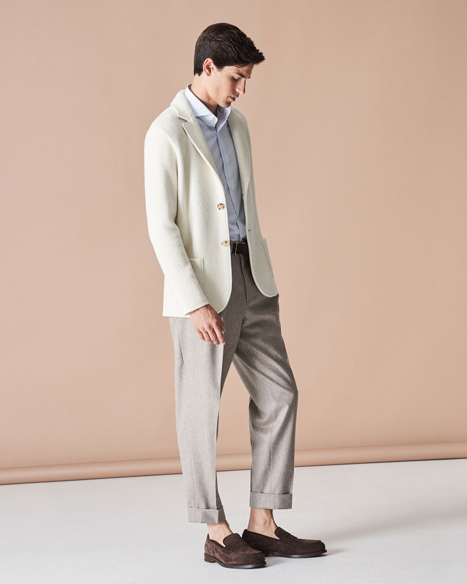 Mann trägt hellblaues Hemd, weißes Sakko, beige Hose mit Umschlag, braunen Gürtel und braune Loafer, steht seitlich. Mann trägt hellblaues Hemd, weißes Sakko, beige Hose mit Umschlag, braunen Gürtel und braune Loafer, steht seitlich.