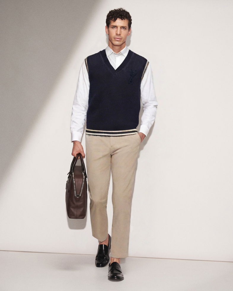 Mann trägt beige Hose, weißes Hemd, dunkelblauen Pullunder, schwarze Loafer und hält braune Ledertasche in der Hand.