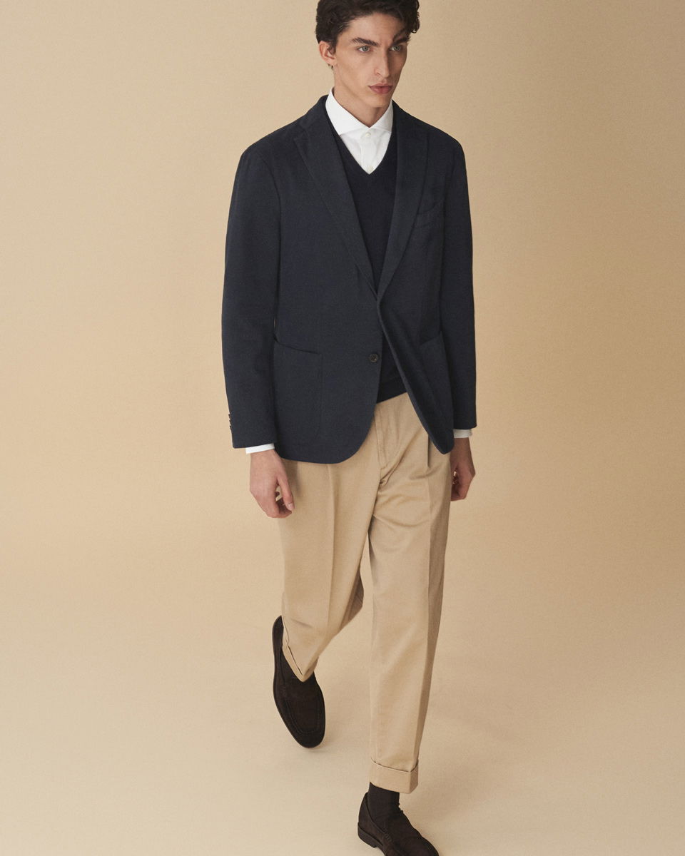 Mann trägt weiße Hemd, schwarzen Pullunder, navy Sakko, beige Hose und braune Loafer, vor beigem Hintergrund.