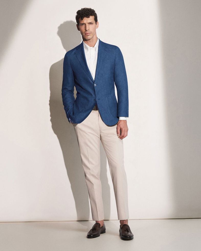 Mann mit blauem Sakko, weißem Hemd, beiger Hose und dunkelbraunen Loafern steht vor hellen Hintergrund.