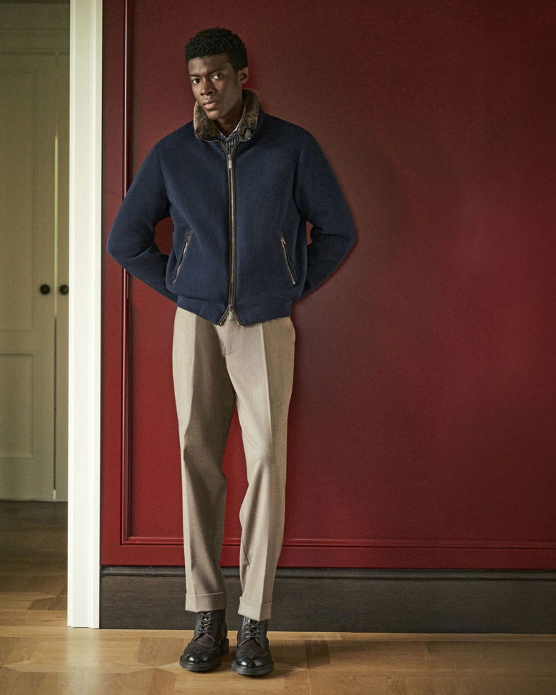 Mann steht vor roter Wand, trägt dunkelblaue Jacke mit Fellkragen, beige Hose und dunkelbraune Schnürschuhe.