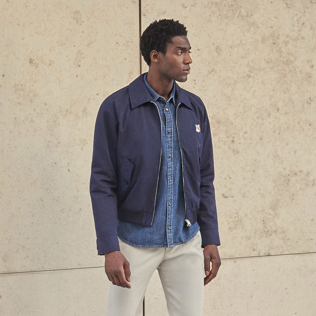 Mann trägt beige Hose, blaues Jeanshemd und dunkelblauen Blouson mit kleinem Logoprint, steht vor heller Wand. Mann trägt beige Hose, blaues Jeanshemd und dunkelblauen Blouson mit kleinem Logoprint, steht vor heller Wand.